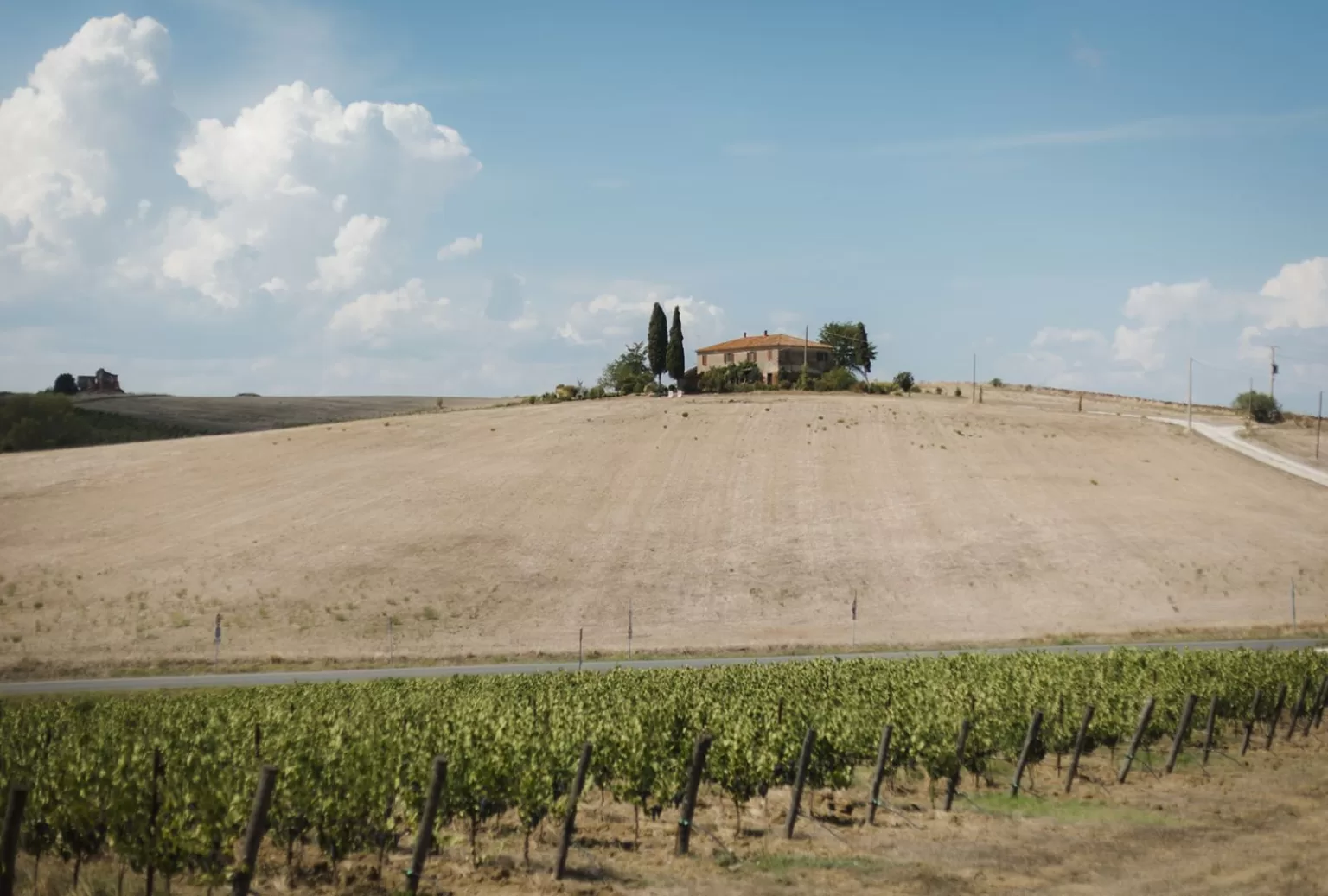A Tuscan farmhouse sits atop a sunlit hill at Borgo Tre Rose, surrounded by rolling fields under a bright blue sky.