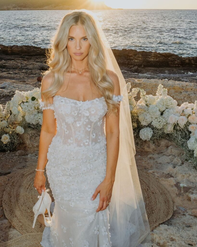 The bride stands barefoot on the rocks, holding her shoes and looking radiant in the evening sun.