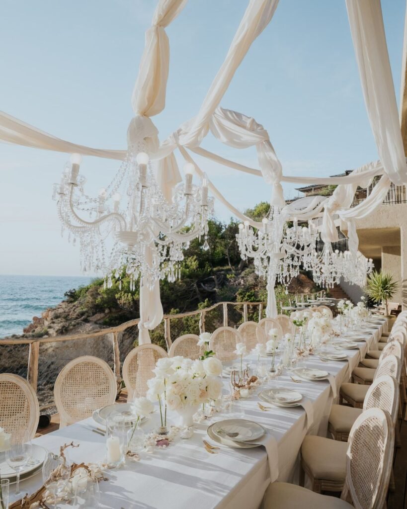 Crystal chandeliers draped with white fabric above an alfresco dinner table set on a wooden deck by the ocean.