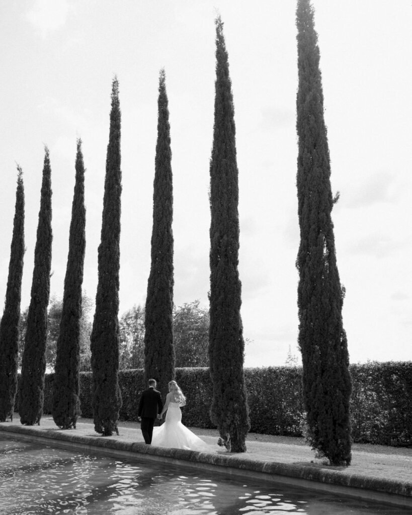 The couple stands between tall cypress trees, their romantic embrace framed by the Tuscan-inspired landscape of Domaine d'Essendieras.