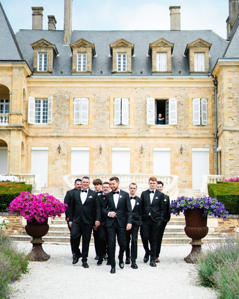 Stylish groom and groomsmen in tuxedos walking confidently outside the elegant Domaine d'Essendiéras château.