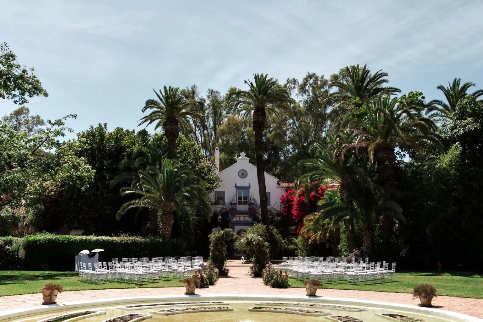 Outdoor wedding ceremony set up in the gardens of Finca La Concepción, with palm trees, white chairs and a historic façade backdrop.