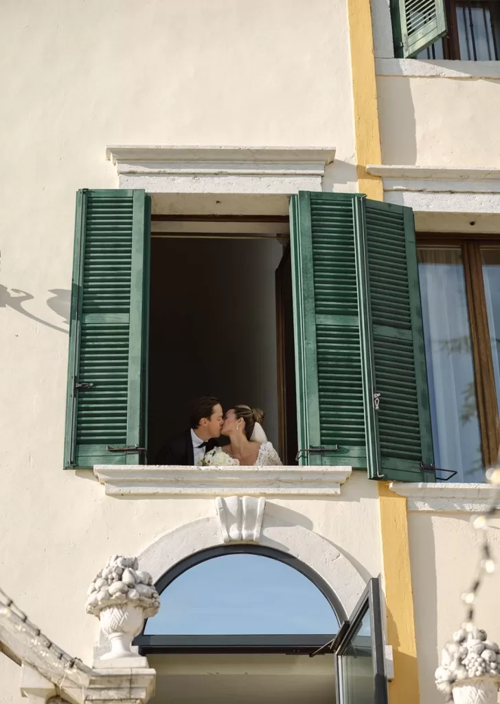 Bride and groom share a kiss at the window of Villa Cariolla, framed by green shutters and golden sunlight.