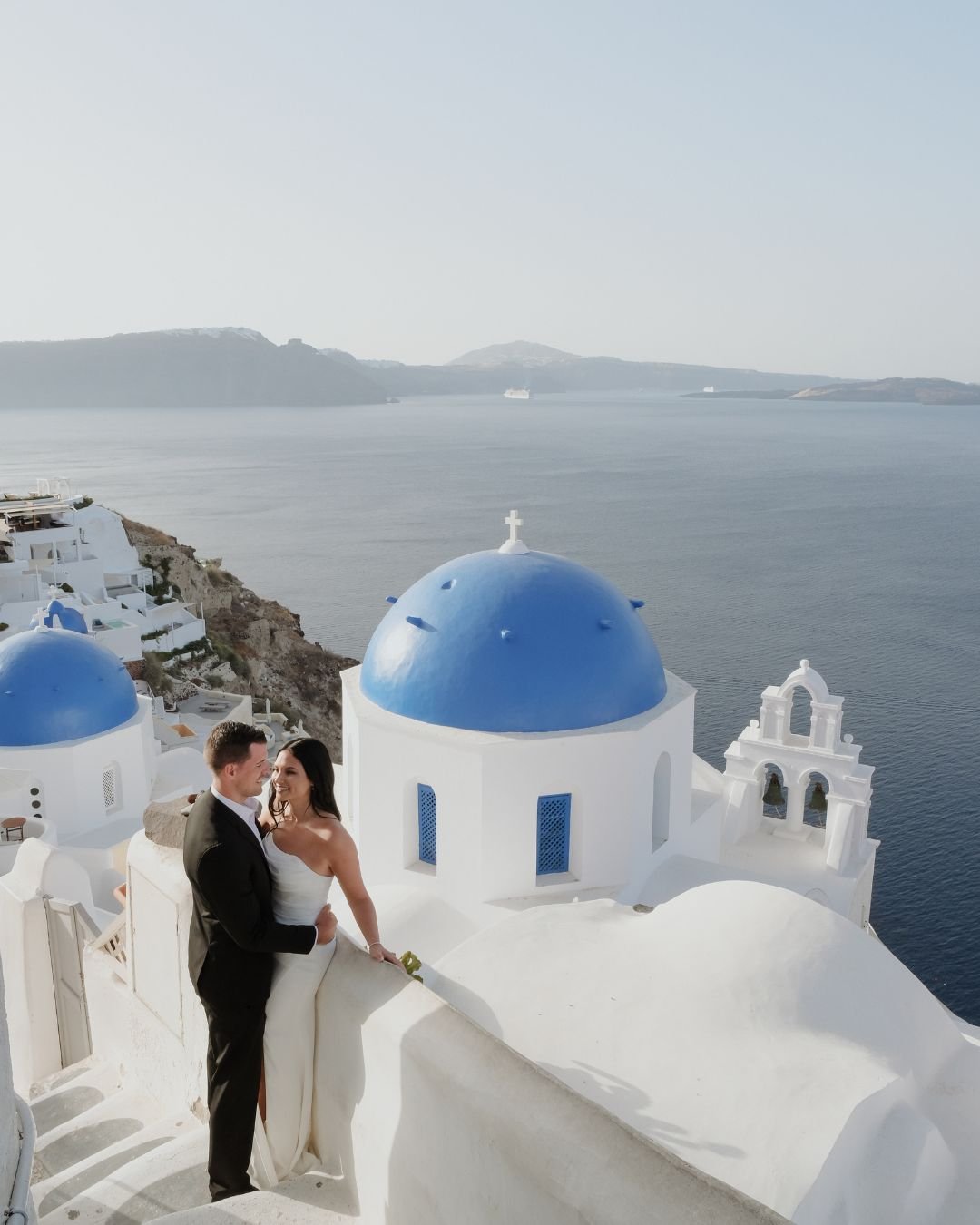 Bride and groom standing on a cliffside in Greece, overlooking the Aegean Sea.