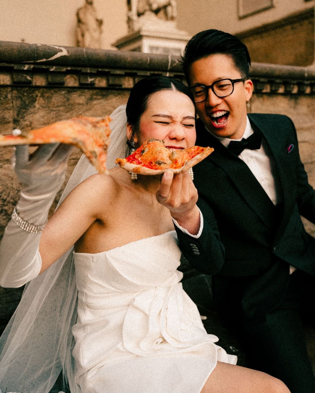 A newlywed couple sharing a slice of pizza in their wedding attire.