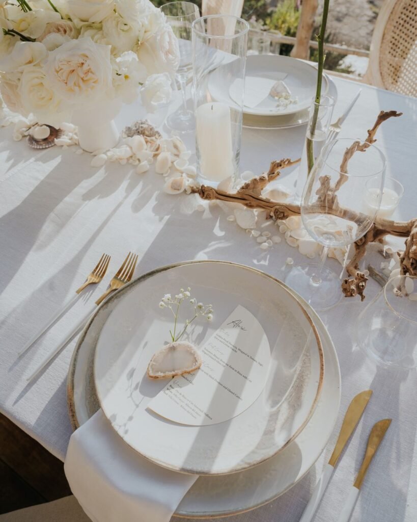 Elegant beachside table setting with a cream-toned plate, shell place marker, and a delicate floral menu under golden hour light.