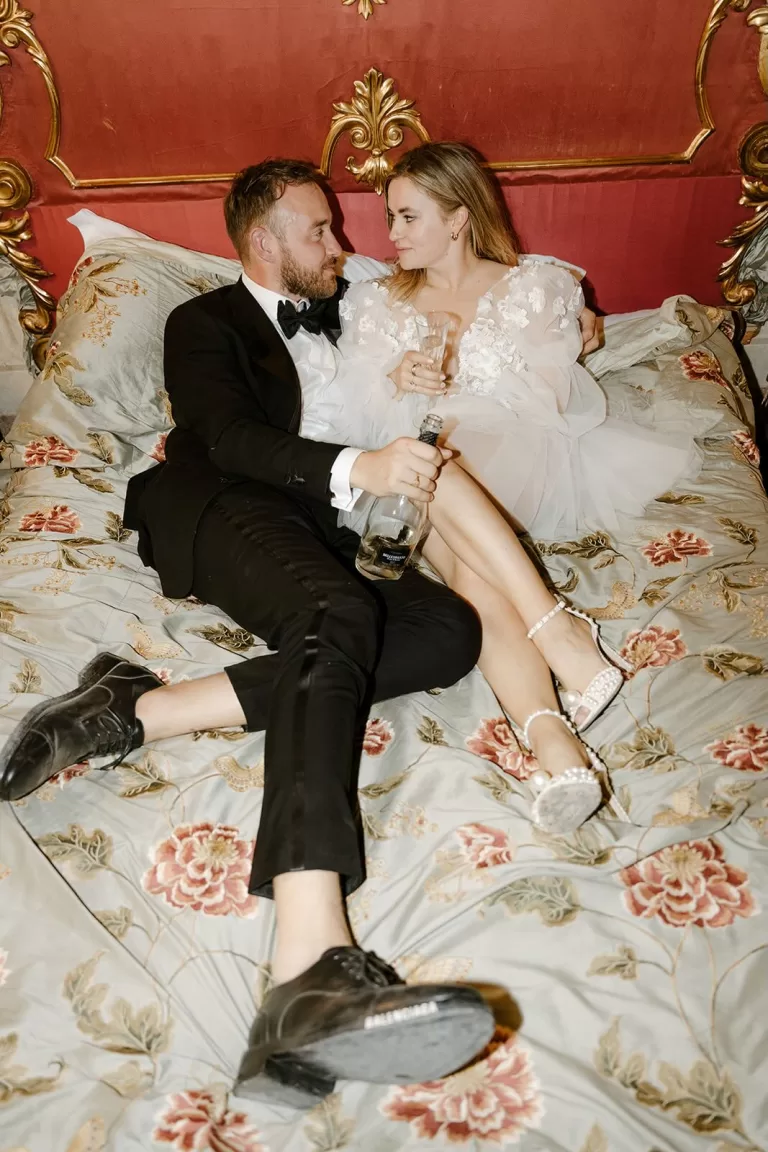 Newlyweds lounging in vintage-style bed at Borgo Stomennano, sharing a quiet moment with champagne.