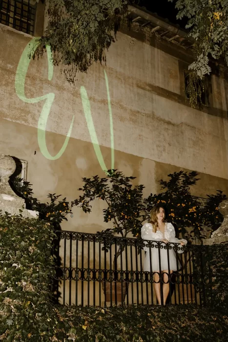 Bride in short tulle dress standing on ivy-draped balcony at Borgo Stomennano, illuminated by monogram projection.