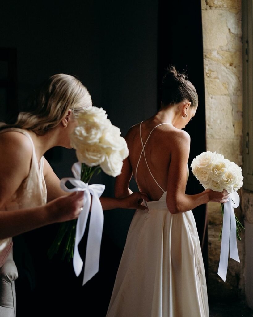 Bride and bridesmaid adjust the dress and hold matching white bouquets tied with ribbon inside Château Rigaud.