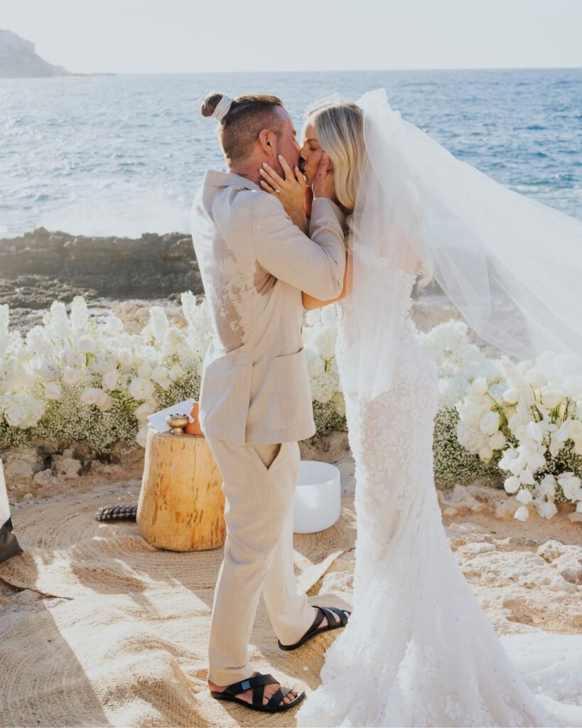 The bride and groom share a kiss after their vows, standing on a clifftop ceremony setup with white florals and the sea behind them.