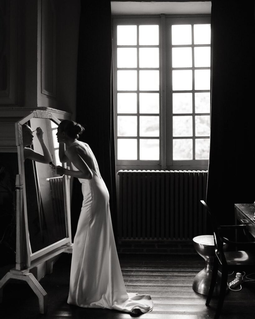 Bride applying makeup at an antique mirror near a tall window at Château Rigaud, captured in quiet focus.