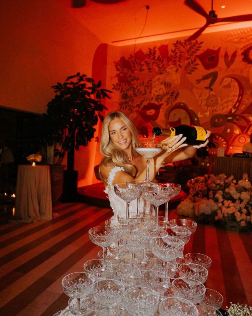 The bride joyfully pours champagne into a tower of coupe glasses during the wedding celebration.