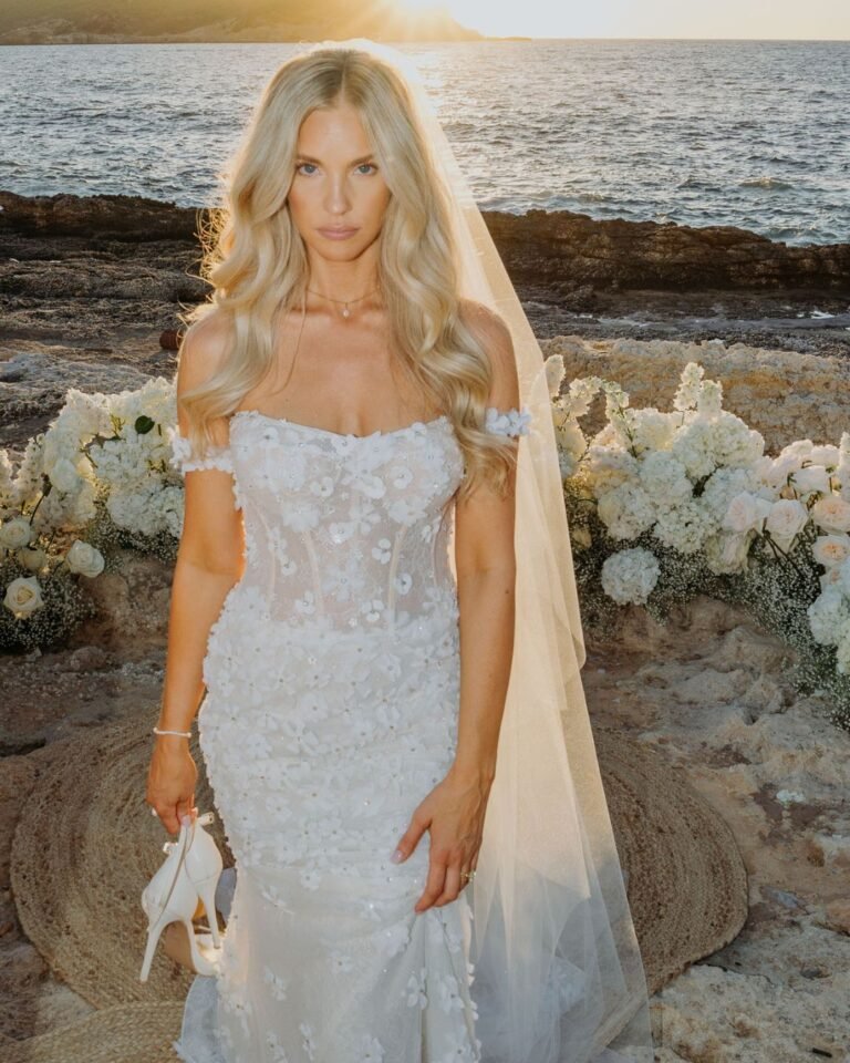 The bride stands barefoot on the rocks, holding her shoes and looking radiant in the evening sun.