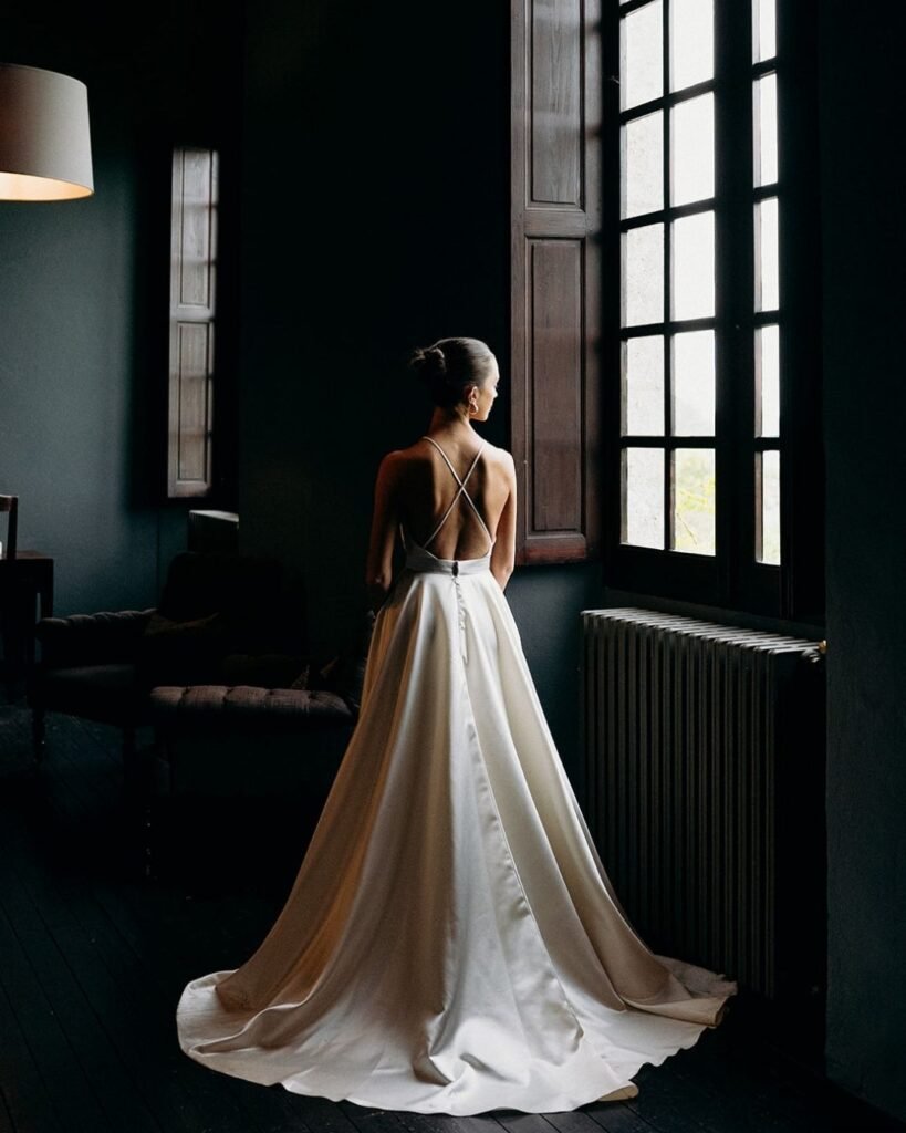 Bride in a minimalist satin gown stands by a large window at Château Rigaud, captured in natural light by Cleya Asulon.