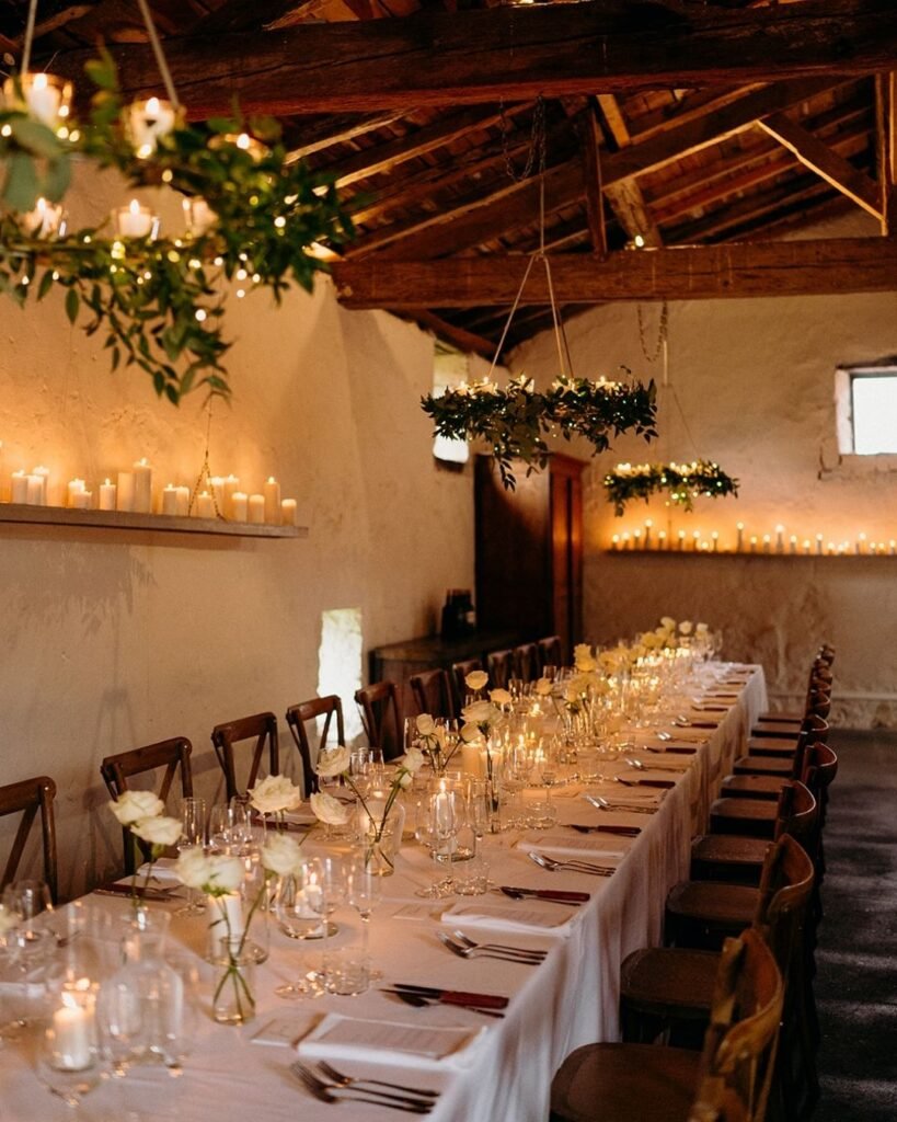 Long candlelit dining table with white roses and soft lighting inside the rustic reception room at Château Rigaud.