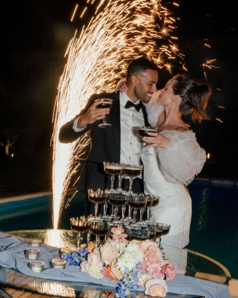 Bride and groom share a kiss during a champagne tower toast with fireworks in the background at Castello Montignano.