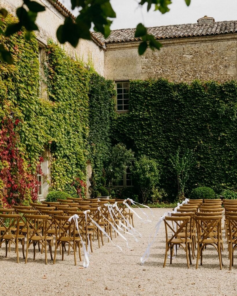 Wooden chairs with white ribbons set for an outdoor wedding ceremony at Château Rigaud, framed by ivy walls.