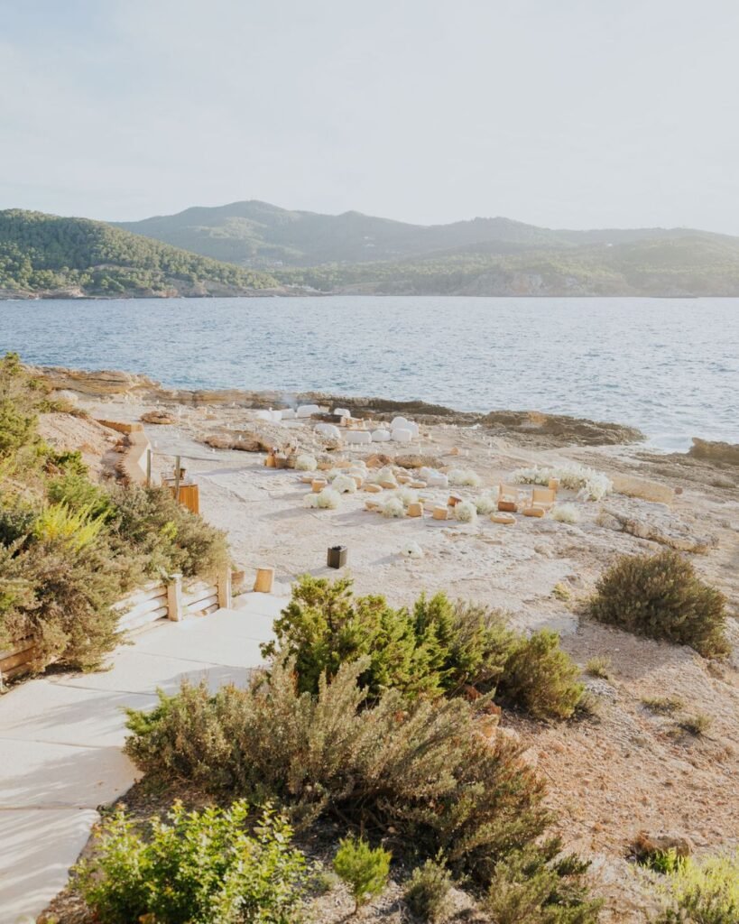 Wide-angle view of the clifftop wedding ceremony site with woven pillows, white florals, and a backdrop of blue sea and green hills.