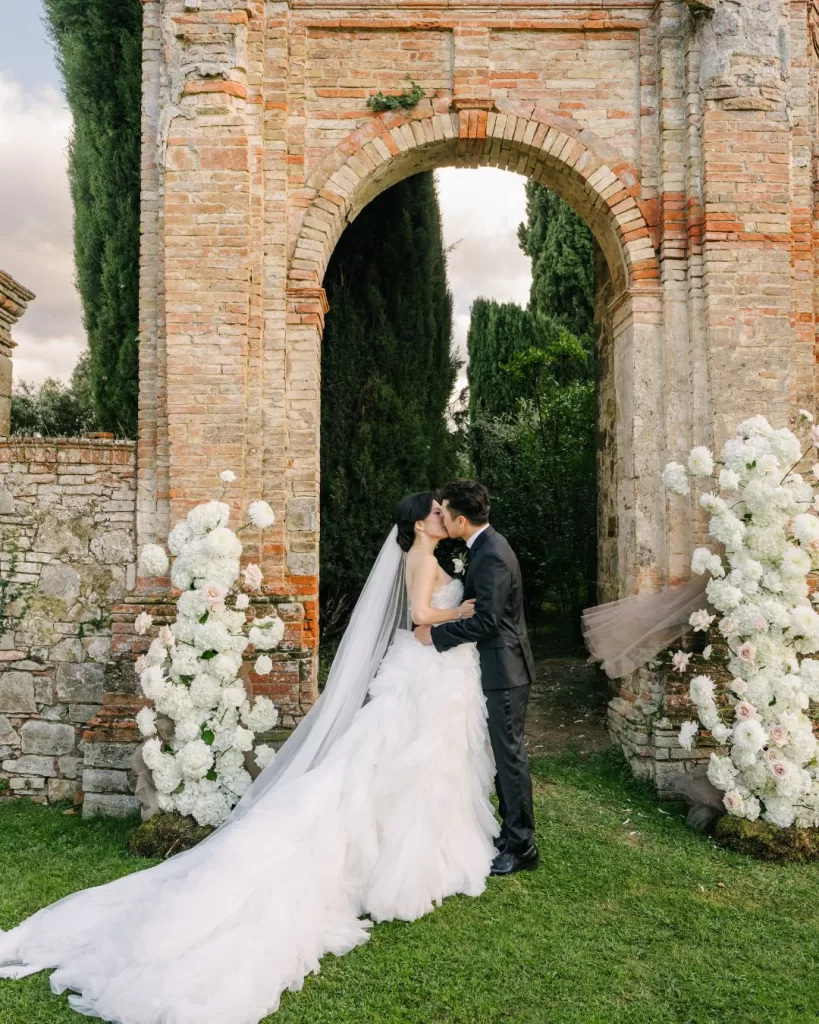 The couple shares their first kiss as newlyweds, framed by the floral ceremony setup at Villa di Geggiano.