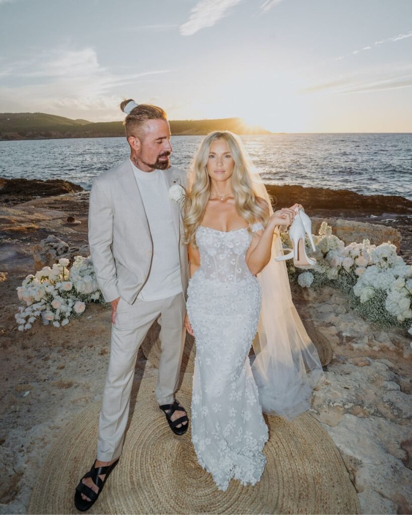 The bride and groom pose by the sea at sunset, the bride holding her heels with a confident expression.