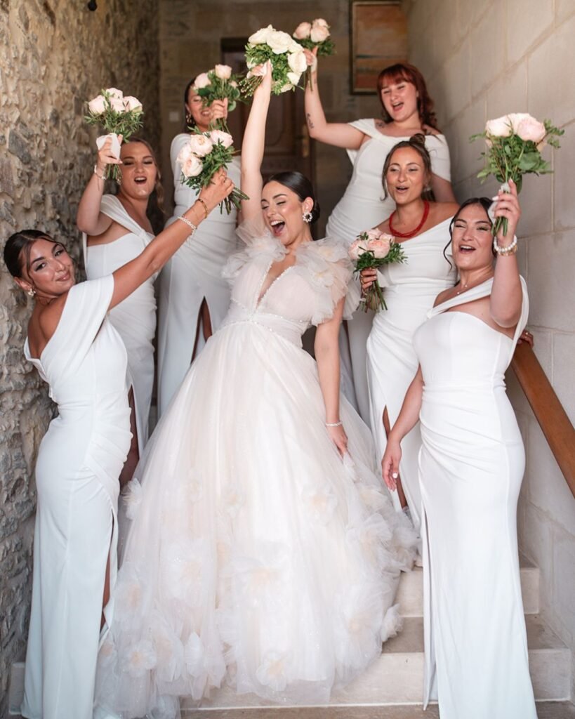 Joyful bride and bridesmaids in white dresses raising bouquets in celebration inside Domaine d'Essendiéras.
