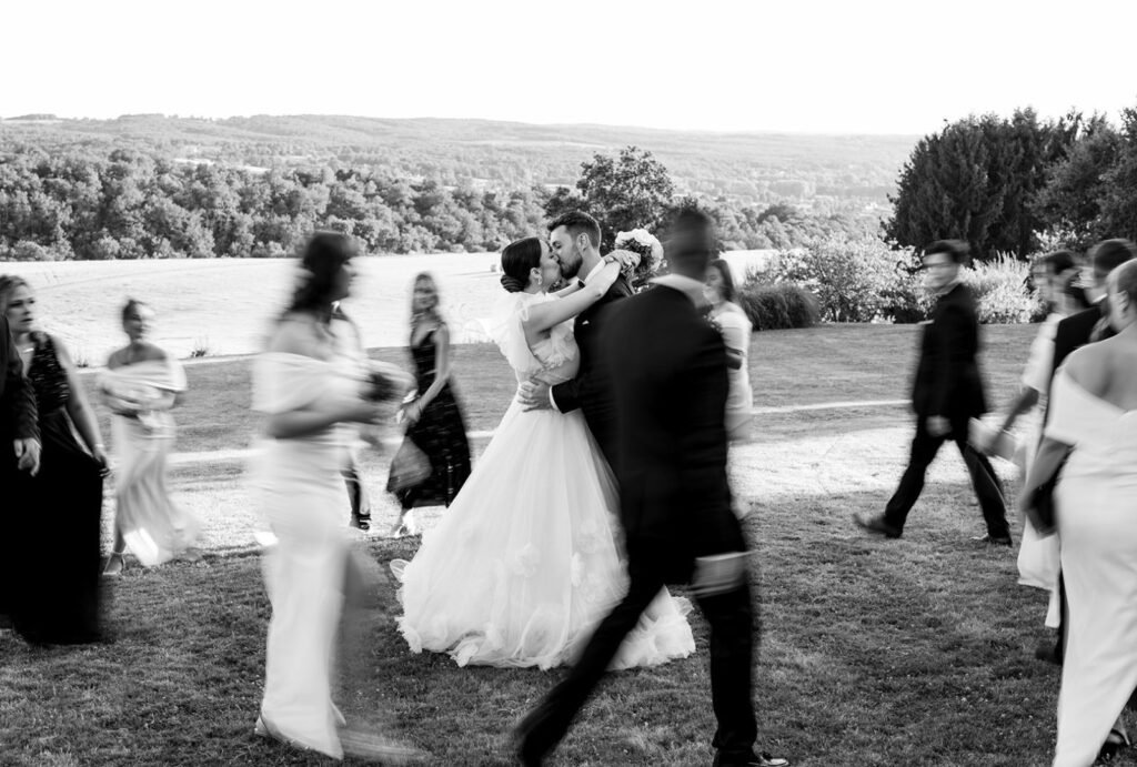 Romantic black-and-white portrait of a bride and groom sharing a kiss, guests moving around them at Domaine d'Essendiéras.