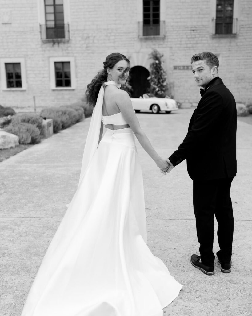 Stylish bride and groom holding hands, looking back over their shoulders toward the camera, with Finca Morneta's impressive facade and wedding car in the background.