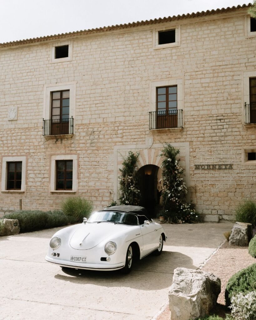 Classic white wedding car parked in front of the historic stone facade and floral-decorated entrance of Finca Morneta.