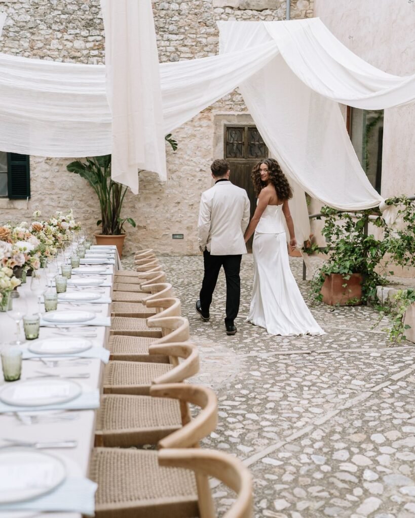 Bride and groom walking hand-in-hand in the charming stone courtyard of Finca Morneta, featuring beautifully styled reception tables.