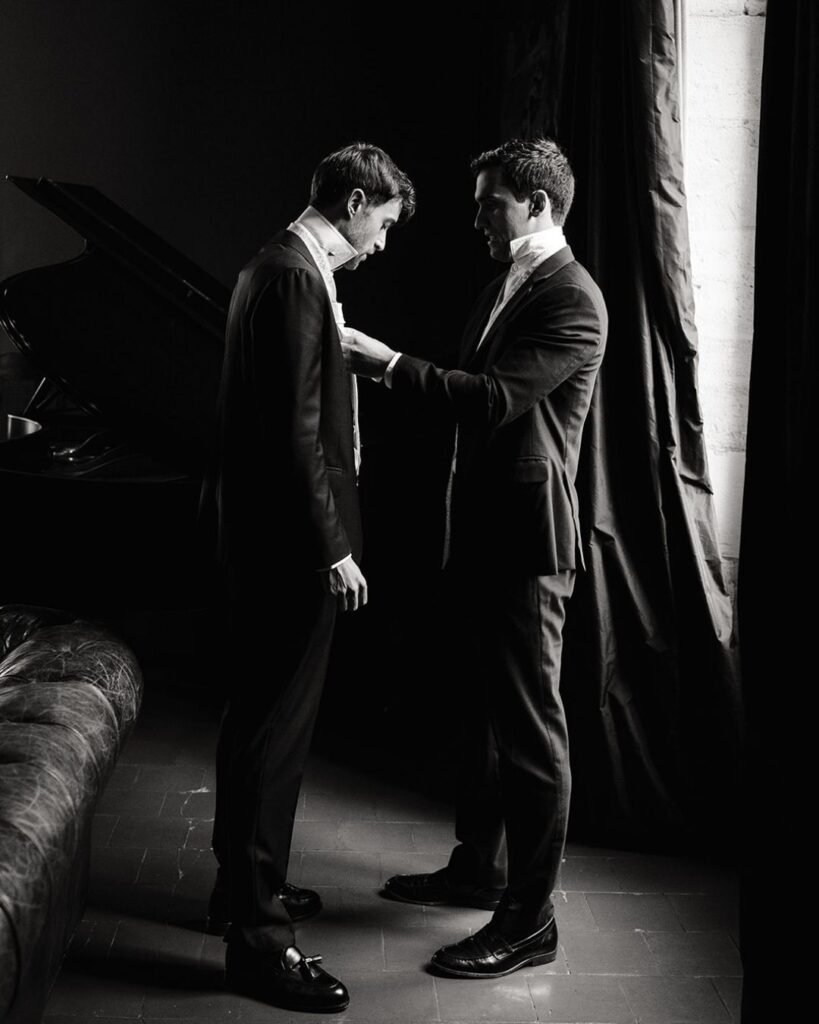 Black and white photo of groomsmen adjusting each other’s suits in soft window light at Château Rigaud.