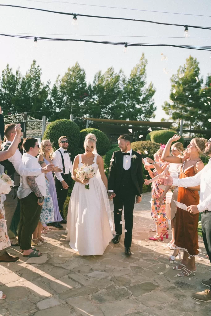 Bride and groom smile at each other, hand in hand, while guests throw petals during their Liopetro celebration.