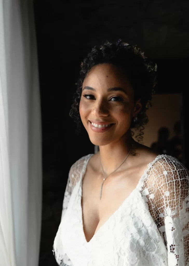 Bride smiling softly by a window in her lace gown at Mas Torroella, with natural light enhancing the serene moment.