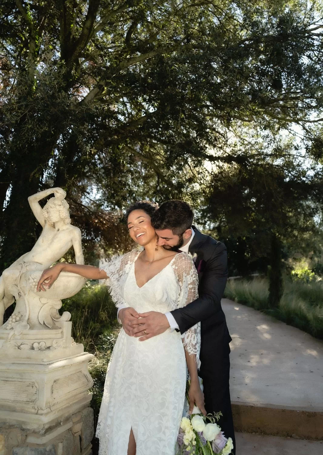 Joyful moment between bride and groom captured by a sculpture in Mas Torroella’s sunlit garden.