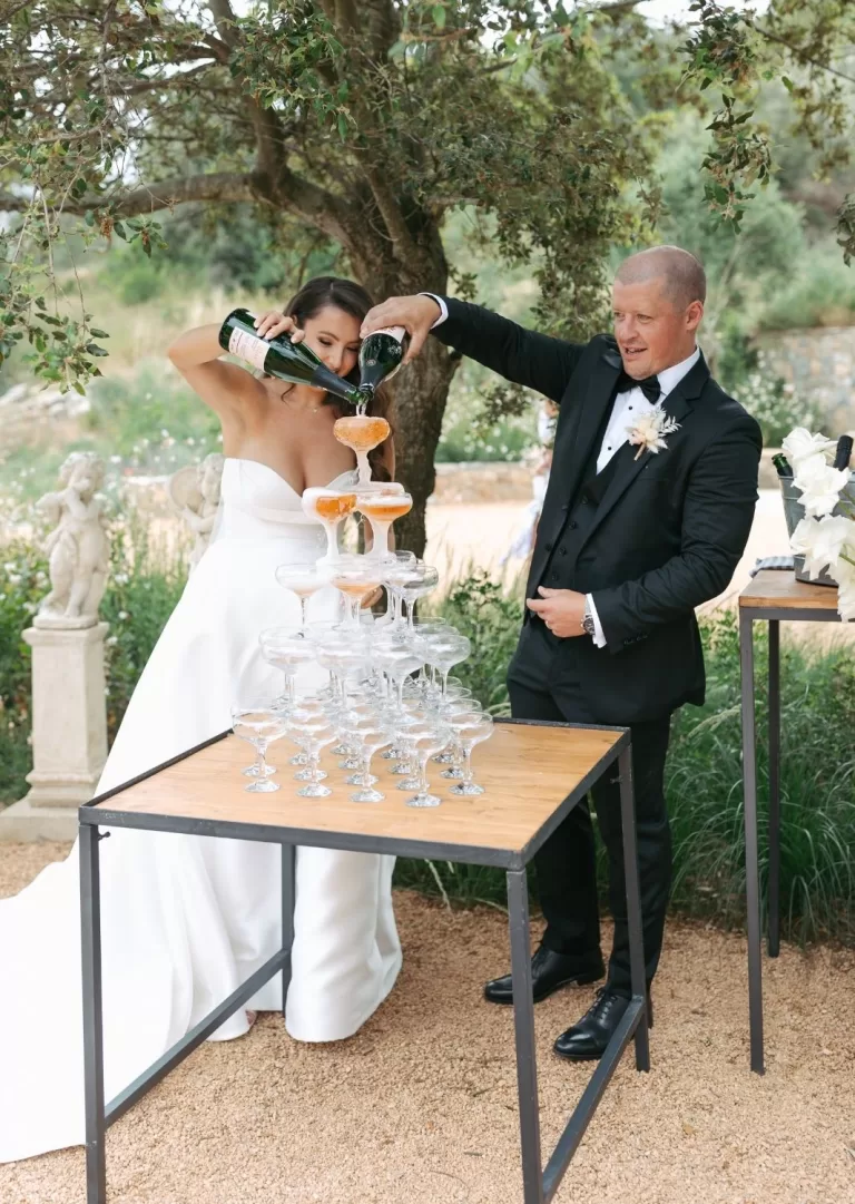 Bride and groom pouring champagne into glass tower under trees at Mas Torroella