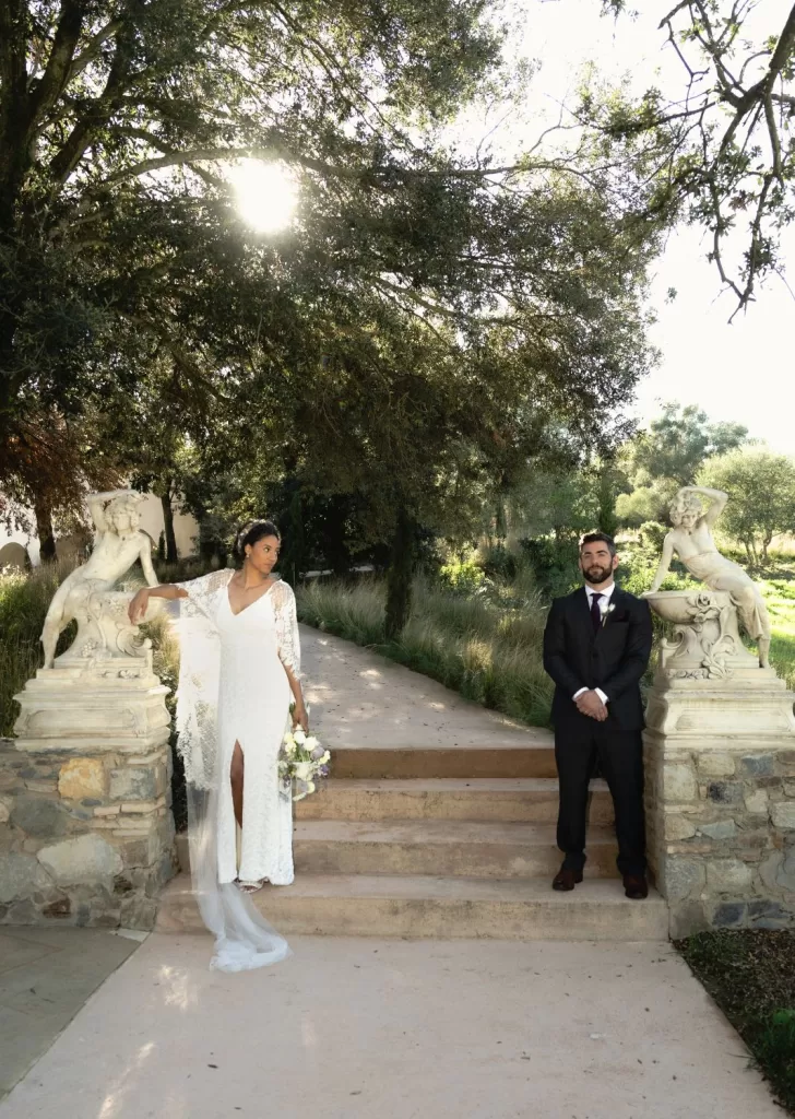 Bride and groom pose near classical statues under dappled sunlight at Mas Torroella