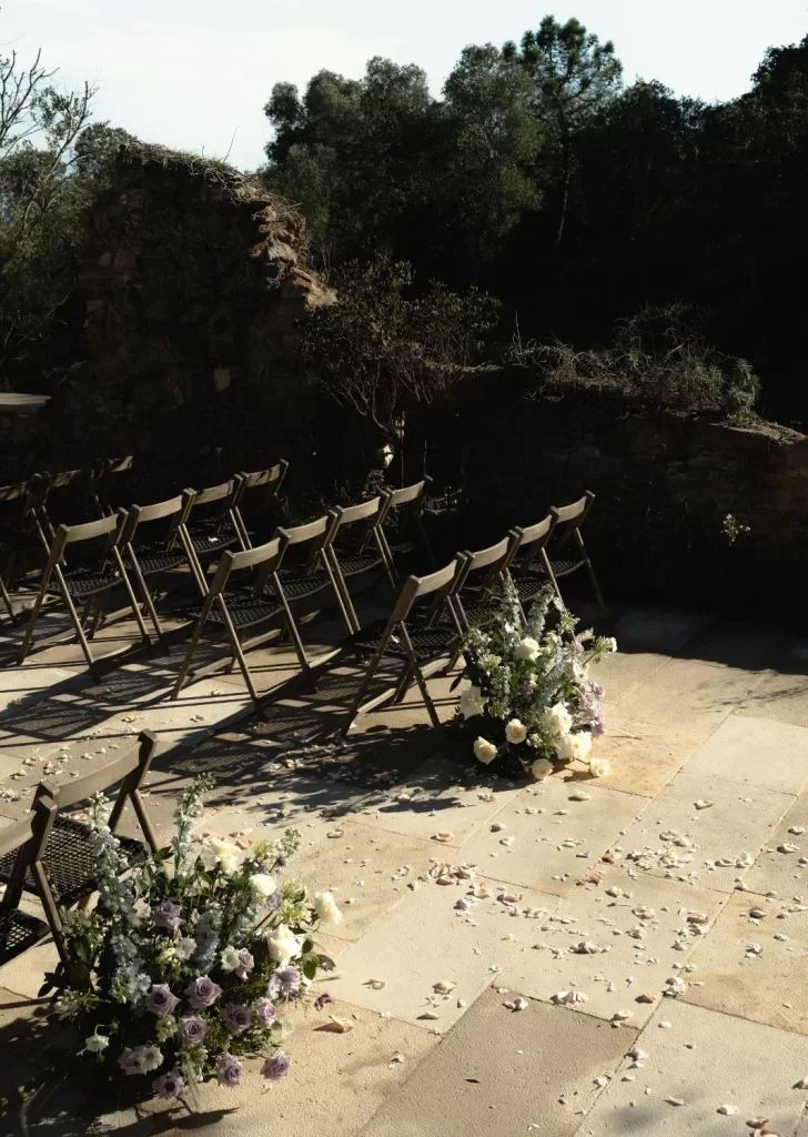 Soft purple and white flower arrangements line the aisle at Mas Torroella, surrounded by natural stone and forest.