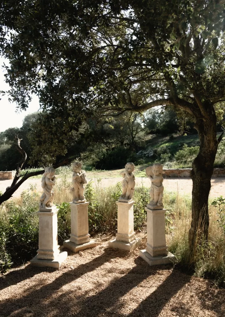 Four cherub statues standing on stone pedestals in a sunlit Mediterranean garden at Mas Torroella.
