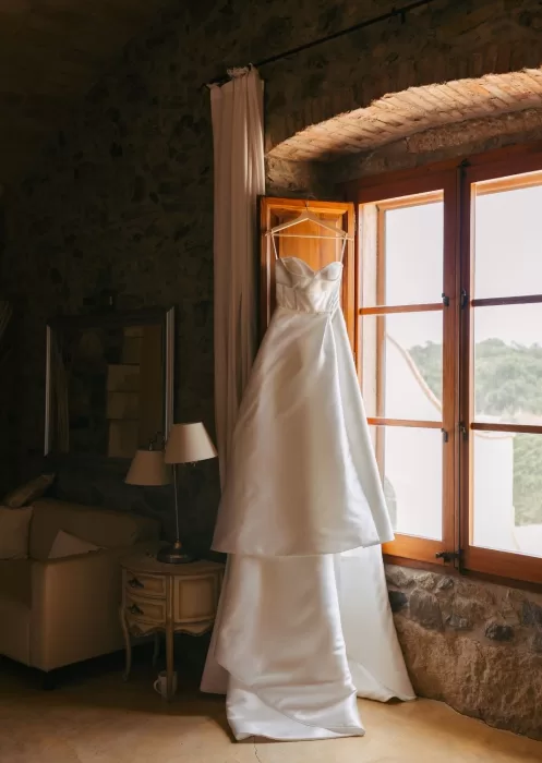 Strapless satin wedding gown hanging in a sunlit stone room window at Mas Torroella.
