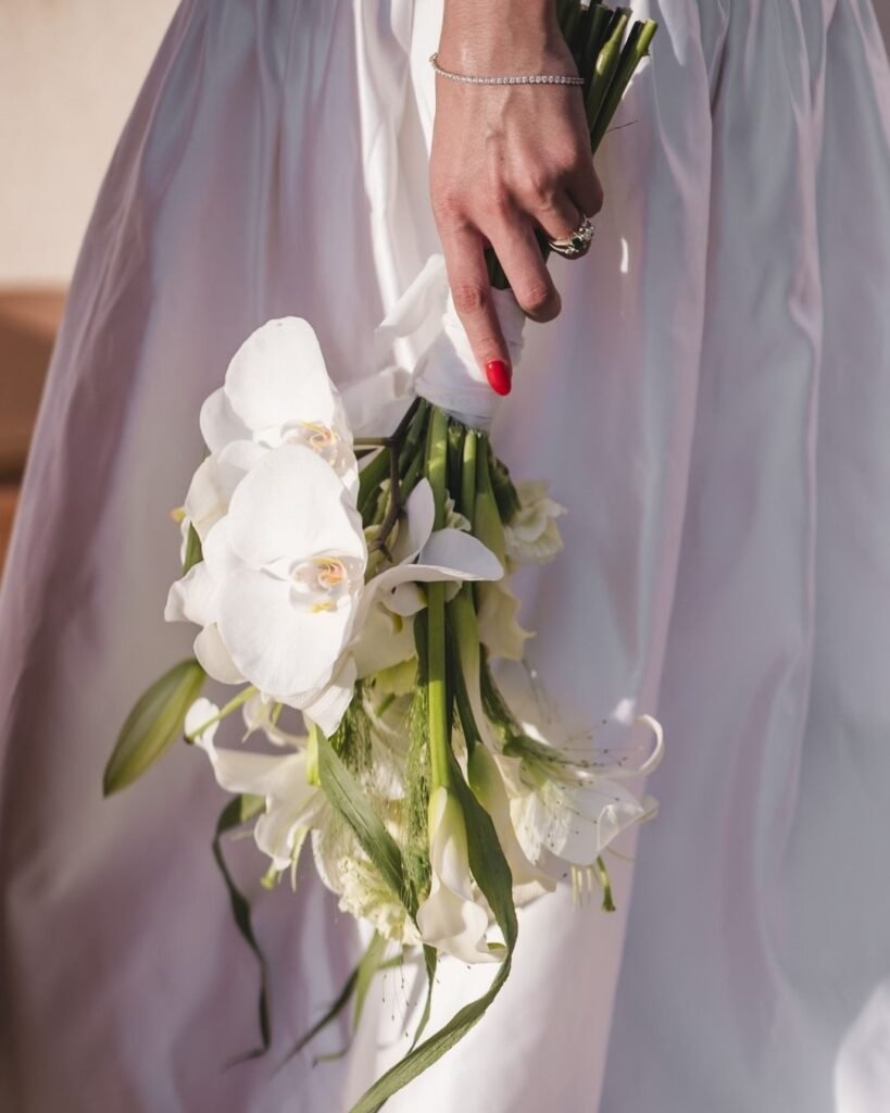 Close-up of bride’s hand holding a cascading white orchid bouquet, with silk dress folds glowing in the sunlight at Masia Cabellut.