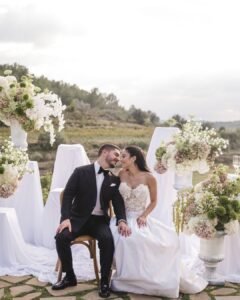 Newlywed couple seated closely, smiling in front of floral arrangements, enjoying their special day at Masia Cabellut.