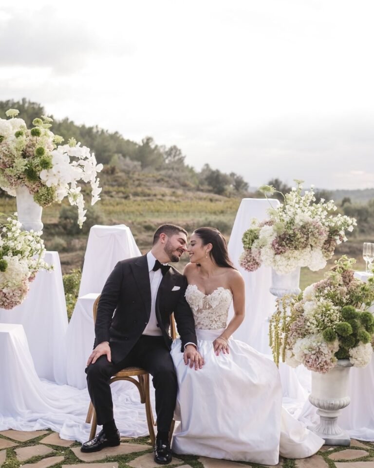 Newlywed couple seated closely, smiling in front of floral arrangements, enjoying their special day at Masia Cabellut.