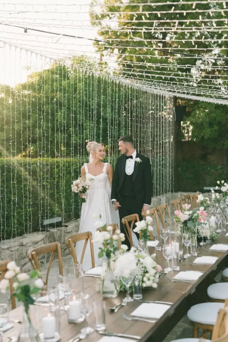 Bride and groom walk hand-in-hand past beautifully decorated reception tables at Liopetro under fairy lights.