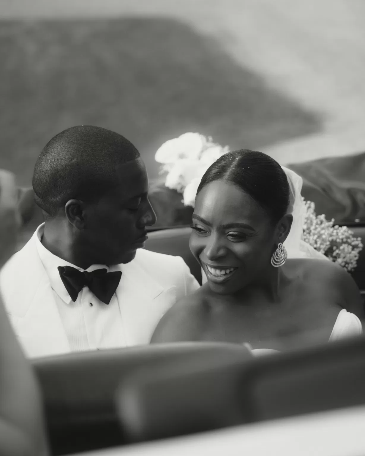 Bride and groom sharing a joyful moment in a vintage car, with the groom in a white tuxedo, captured by Ruben Larruy.