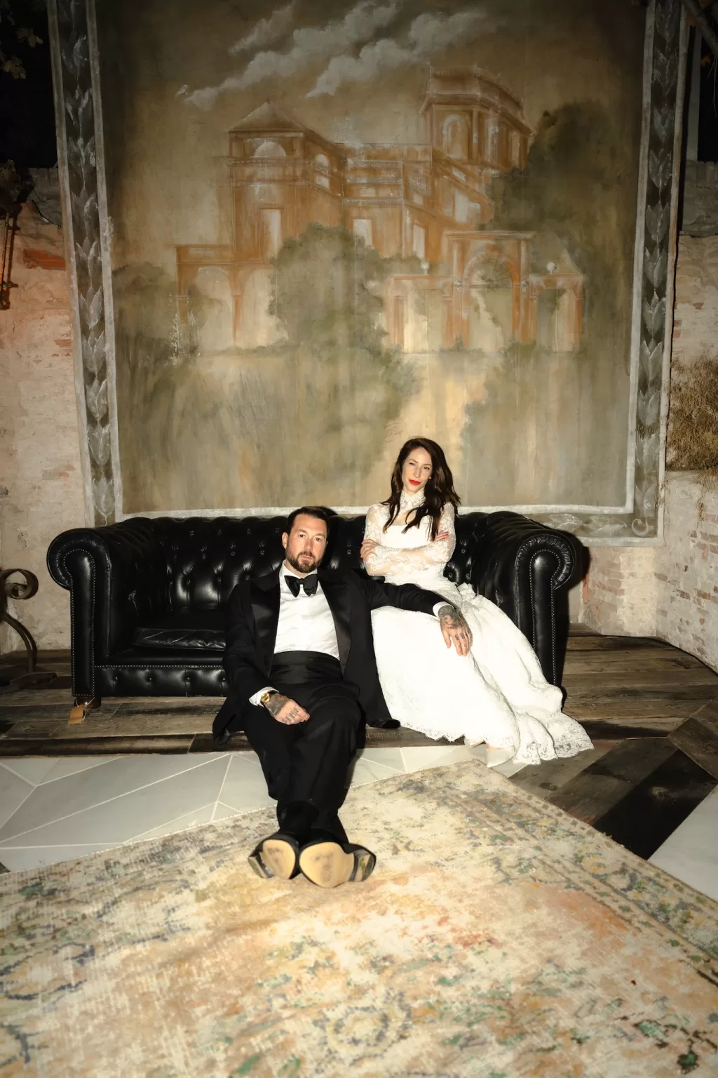 A couple in formal wedding attire sitting on a black leather sofa under a painted mural, intimate and moody portrait by Ruben Larruy.