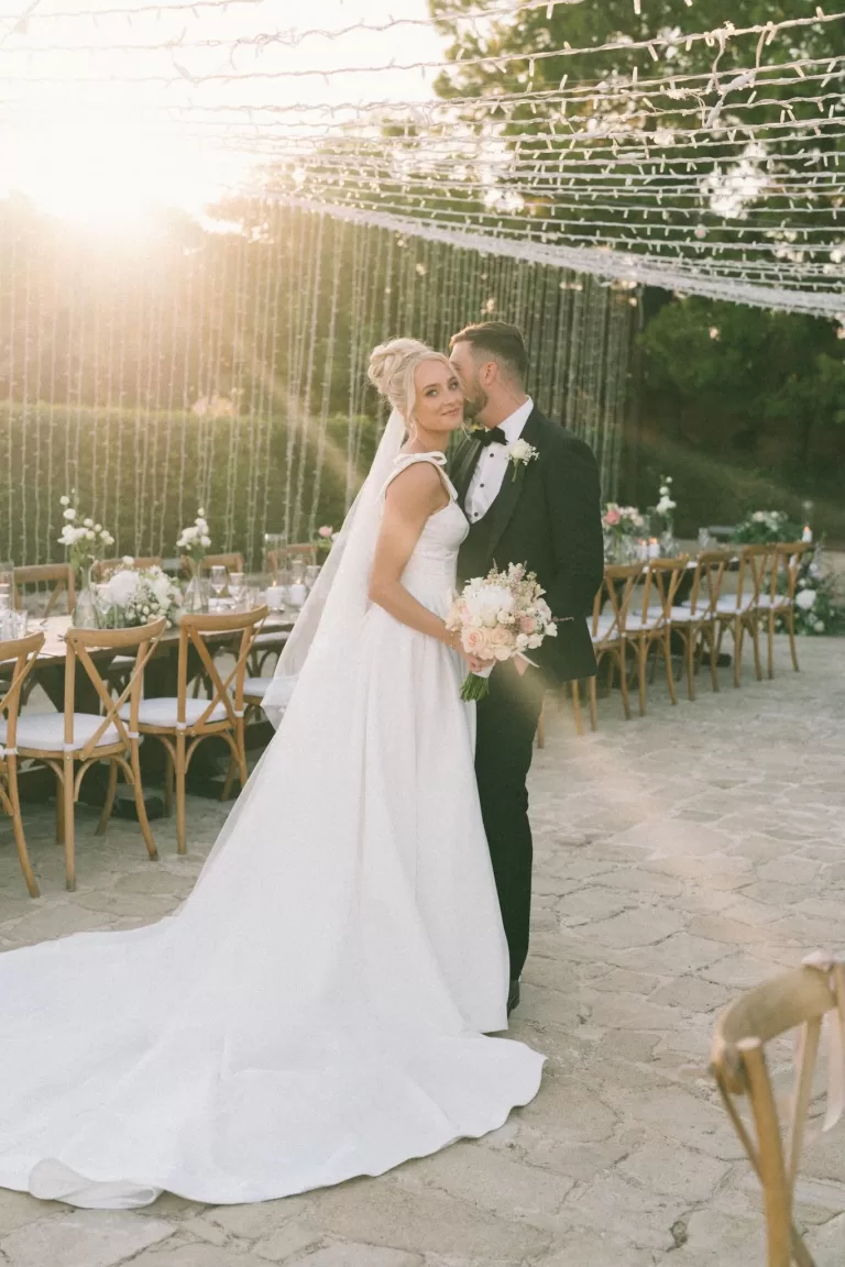 Groom kisses his bride’s temple as she smiles, wrapped in golden-hour light at Liopetro’s outdoor reception.