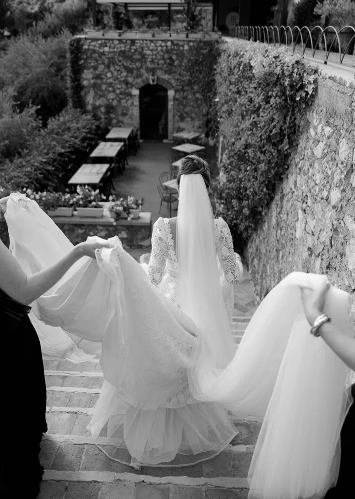 Bride walking down stone steps at Villa Cariola with her lace gown and veil elegantly carried by her bridesmaids.