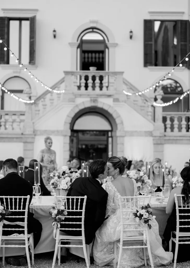 Romantic kiss between bride and groom during their outdoor dinner reception at Villa Cariola, with string lights overhead.