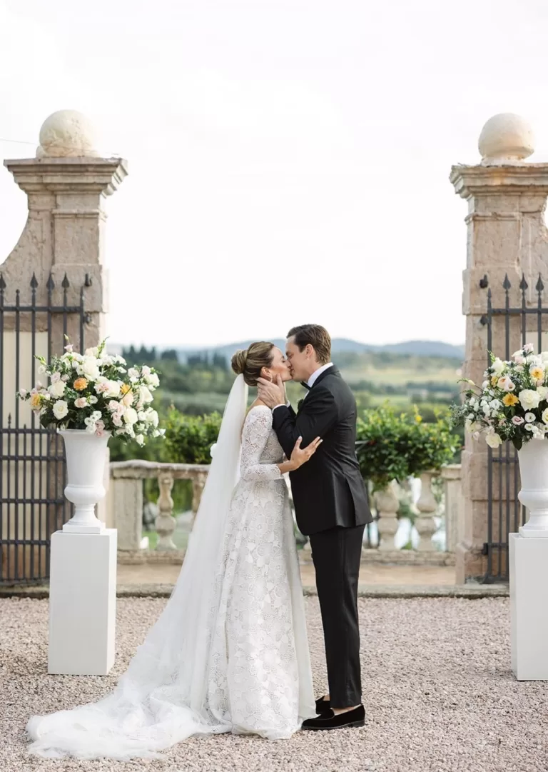 Bride and groom kiss at the altar framed by floral urns and stone pillars at Villa Cariolla, with rolling hills in the background.