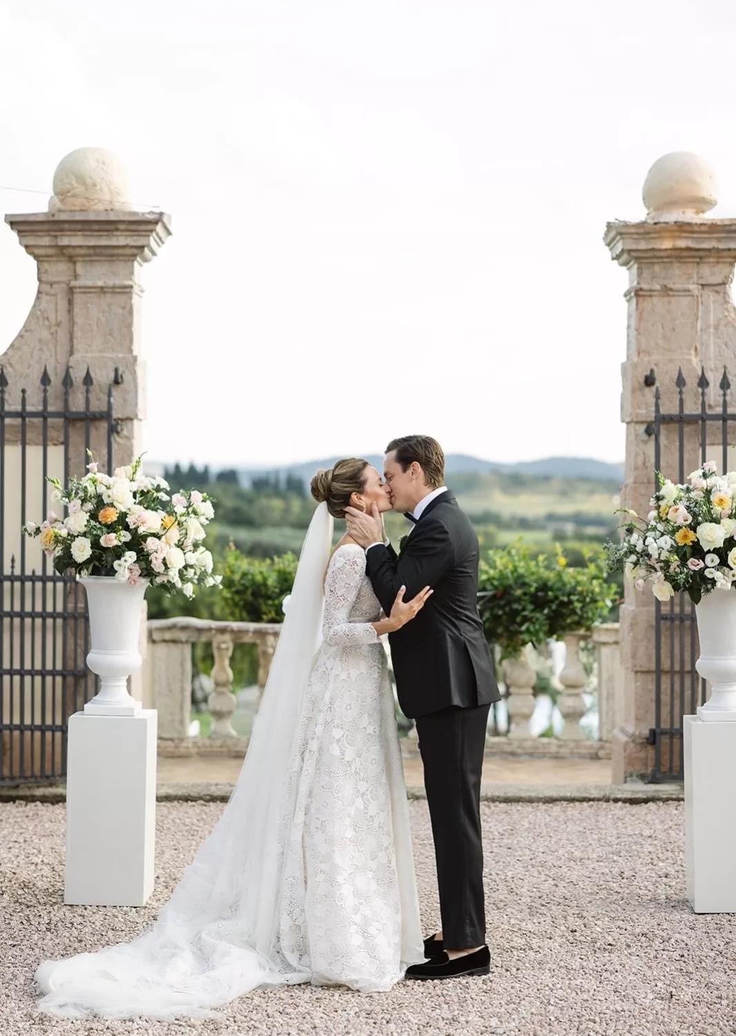 Bride and groom kiss at the altar framed by floral urns and stone pillars at Villa Cariolla, with rolling hills in the background.