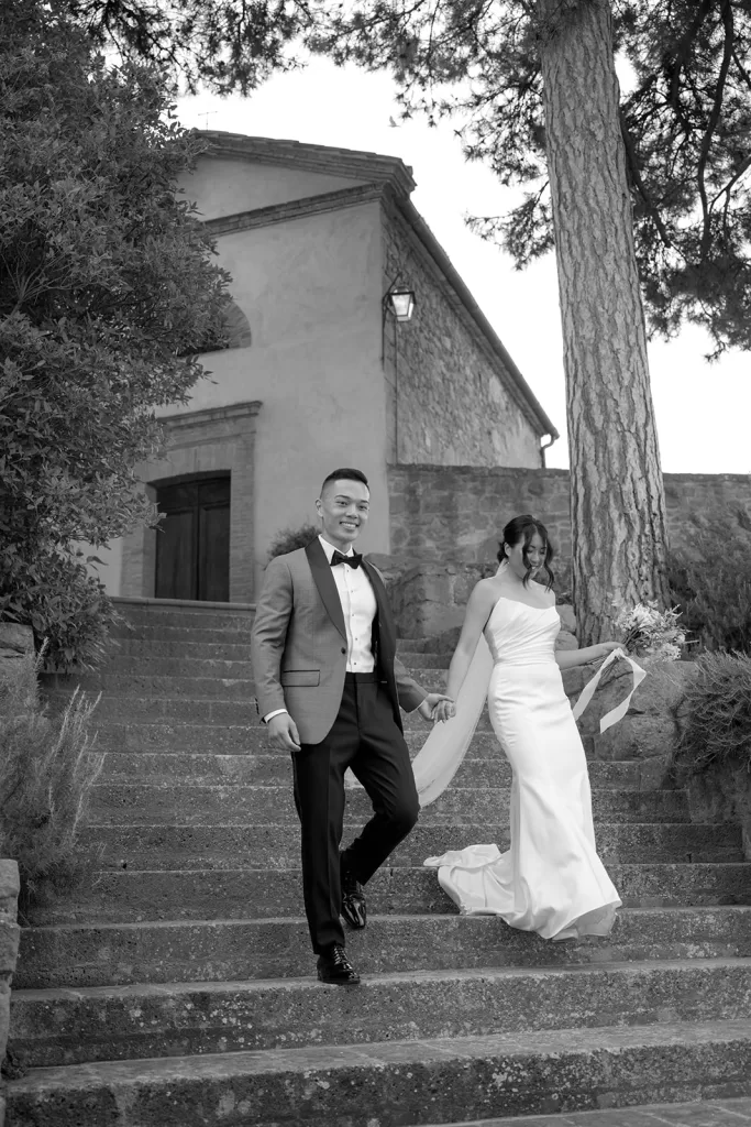 Black and white image of a couple walking down the chapel steps at Borgo Sant Ambrogio, surrounded by stone and pine trees.